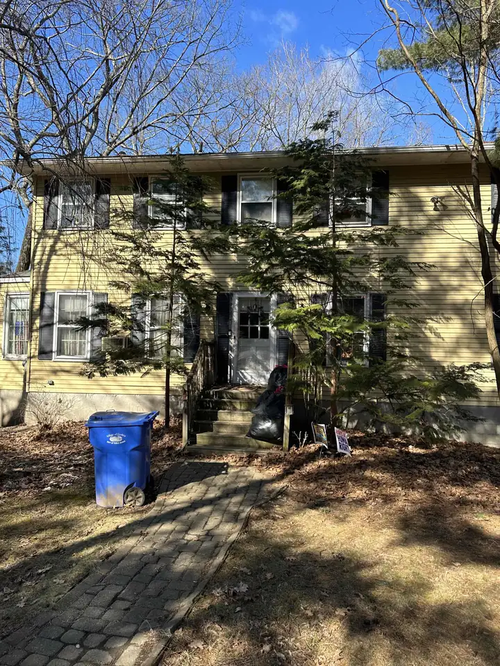 A yellow two-story colonial house with black shutters, a brick paver walkway and bare winter trees
